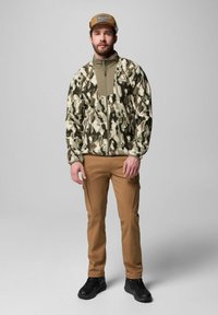 Tarnfarbenes Fleecesakko in Grüntönen, mit Reißverschluss vorne, hohem Kragen; kombiniert mit beigefarbenen Cargo-Hosen und schwarzen Schuhen. Braune Kappe mit Logo.