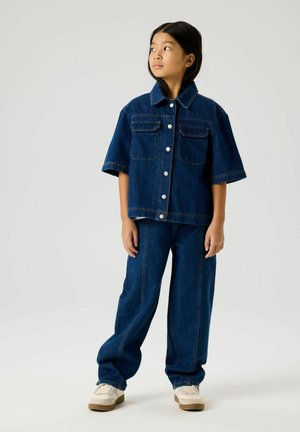 Child standing, wearing dark blue denim jacket with short sleeves, matching loose jeans, and white sneakers, looking to the side.