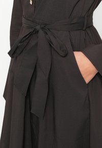 Schwarzes Kleid aus glattem Stoff, mit einem gebundenen Gürtel in der Taille, langen Ärmeln undseitlichen Taschen.