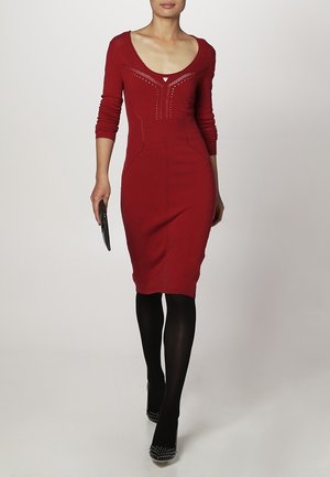Czerwona dzianinowa sukienka z długimi rękawami, głębokim dekoltem w kształcie litery V, dopasowana w sylwetce, z subtelnymi zdobieniami, noszona z czarnymi rajstopami i słupkowymi szpilkami z ćwiekami.