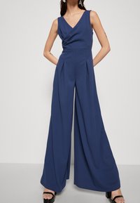 Marineblaue, ärmellose Jumpsuit mit V-Ausschnitt, gewickeltem Oberteil, plissierten weit geschnittenen Beinen und glatter Textur. Kombiniert mit schwarzen Sandalen mit Blockabsatz.