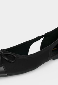 Chaussure de ballet plate noire avec un bout arrondi, petit détail nœud et sangles latérales élastiques pour un maintien sûr.