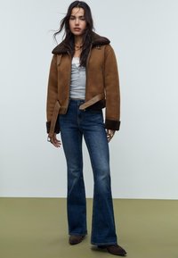 Veste en shearling marron avec une sangle en cuir marron, chemise grise en dessous, jeans évasés bleus et bottines à chevilles marron foncé.