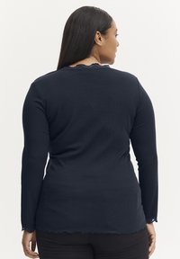Marineblå langærmet top med ribbet tekstur, scalloped blondeudskæring og en let ujævn kant. Båret med sorte bukser, set fra ryggen.