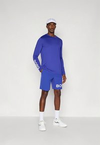 Blue long-sleeve top with "BORC" printed on the sleeve, paired with matching shorts featuring "BORC" on the side. White sneakers and cap.