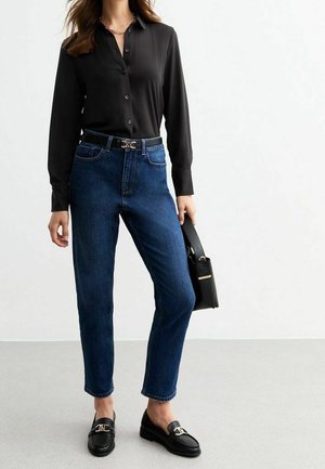 Vrouw die een zwarte blouse met knopen draagt, blauwe high-waist jeans, zwarte loafers en een zwarte handtas met gouden accenten vasthoudt.