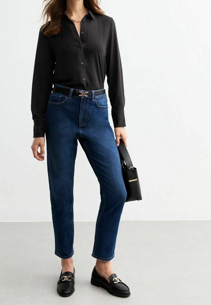 Femme portant un chemisier noir boutonné, un jean bleu taille haute, des mocassins noirs, et tenant un sac à main noir avec des accents dorés.