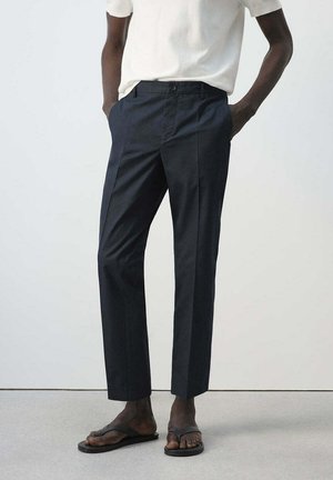 Persona con pantalones negros ajustados y chanclas negras, con las manos en los bolsillos y una camiseta blanca de manga corta sobre un fondo liso.