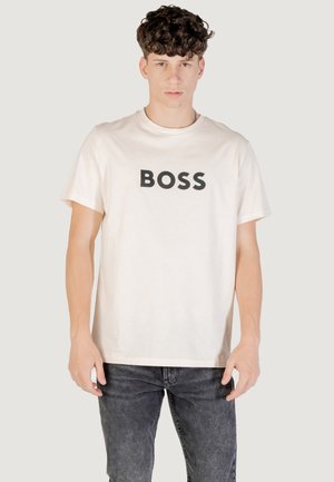 Jonge man met krullend haar die een wit T-shirt draagt met 'BOSS' in zwart gedrukt, gecombineerd met donkergrijze spijkerbroek, staand tegen een lichte achtergrond.