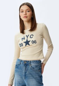 Beige lange mouwen top met geribbeld textuur. Bevat een blauwe geborduurde "NYC 20 06" en sterrenontwerp. Gecombineerd met lichtblauwe denim jeans.