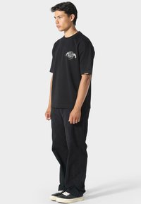 Sort bomulds t-shirt med korte ærmer, der har et grafisk design på brystet. Sammen med løstsiddende sorte bukser og sneakers.