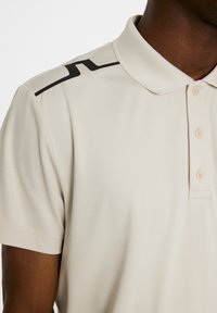 Beige pikétröja med en strukturerad tyg, med en tvåknapps knäppning och svarta detaljer på axeln.