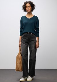 Zelený pletený svetr s výstřihem do V, černé široké kalhoty, bílé tenisky a béžová semišová taška. Jednoduchý, moderní design s volným střihem.