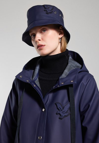 Marineblaue wasserdichte Jacke mit Kapuze und Adler-Logo, kombiniert mit einem passenden Bucket Hat. Darunter trägt sie einen strukturierten schwarzen Rollkragenpullover.