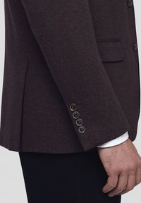 Burgundowa teksturowana marynarka z czterema guzikami na mankietach, pojedynczą kieszenią na piersi i rozcięciem bocznym. Tkanina wydaje się pleciona i oddychająca.