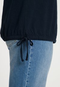 Marineblauwe top met verzamelde elastische zoom en verstelbare trekkoord, gedragen over lichtblauwe denim jeans, met nadruk op de zachte stofstructuur.