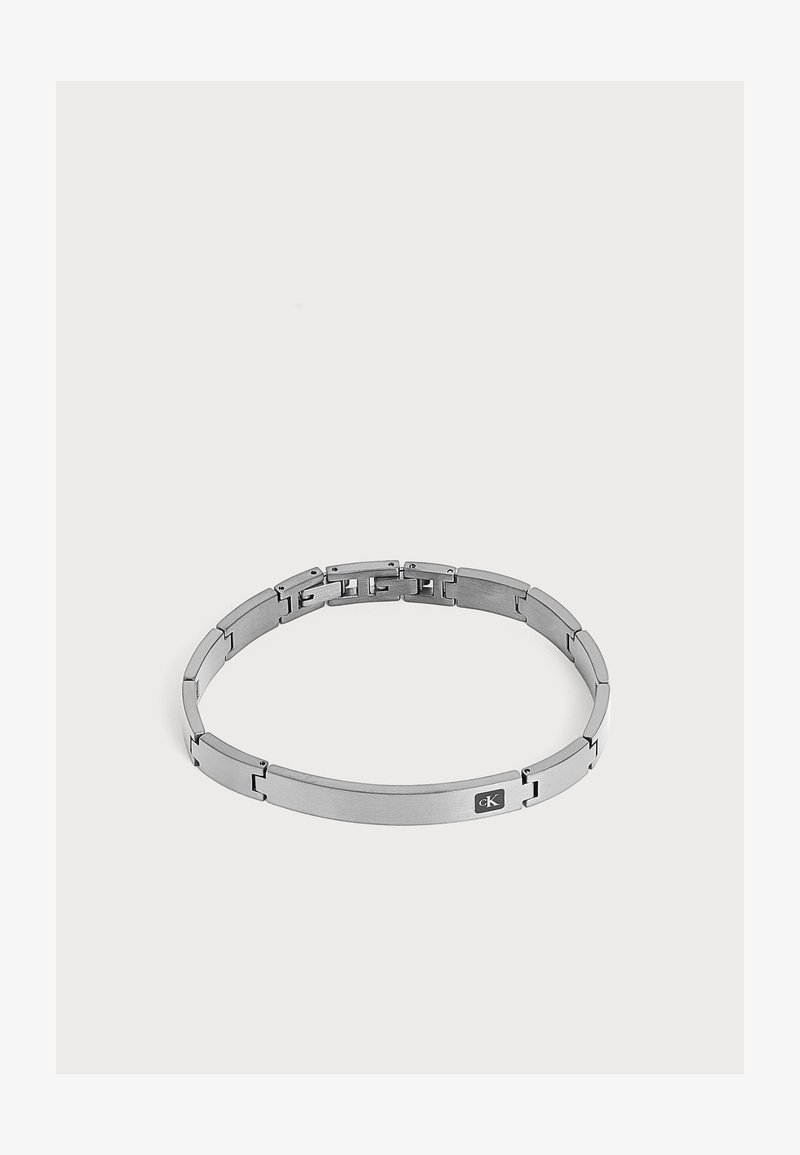 Bracelet en métal argenté avec une plaque rectangulaire comportant un petit logo "cK" sur un côté.
