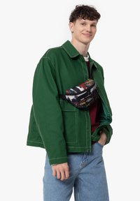 Zelená plátěná bunda s bílým prošíváním, která je nošena přes burgundské tričko, s černým bederním batohem s barevnými graffiti vzory.