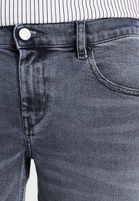 Vaqueros de mezclilla gris con un diseño clásico de cinco bolsillos, cierre frontal con botón y una sutil textura con un acabado ligeramente desgastado.
