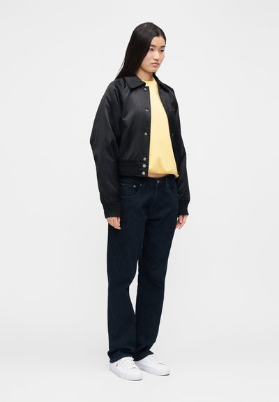 Čierna saténová bomber bunda so zapínaním na cvoky cez žlté tričko s okrúhlym výstrihom, doplnená tmavomodrými džínsami a bielymi teniskami.