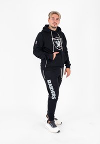Zwarte hoodie met Raiders-logo, witte koordjes; zwarte joggers met "RAIDERS" in zilver gedrukt. Draag sneakers in wit en zwart.