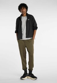 Musta kevyt takki, harmaa t-paita ja oliivinvihreät jumppahousut ribbaetuilla hihansuilla. Mustat lenkkarit, joissa on vaalea pohja. New Balance -logo näkyvissä.