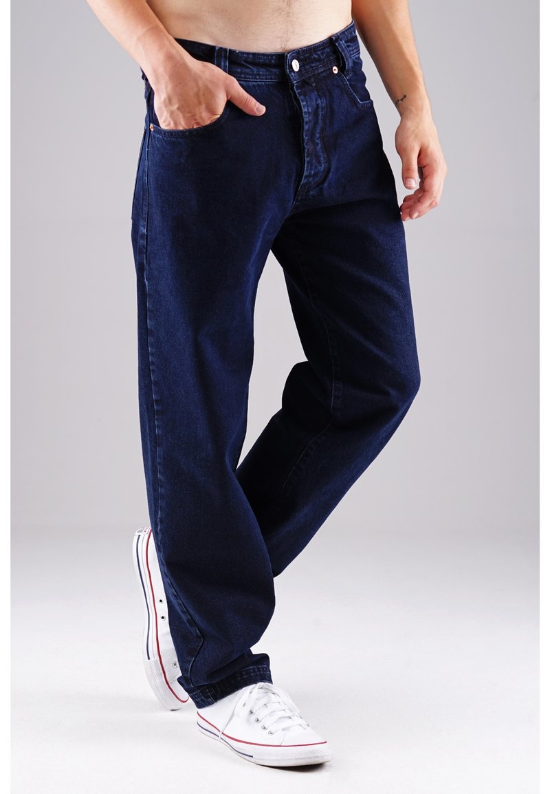 Dunkelblaue Jeans aus Denim mit geradem Schnitt, ausgestattet mit einer Fronttasche und einem Metallknopfverschluss. Getragen mit weißen Sneakern.