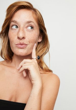Primer plano de una persona con cabello rojo y pecas, usando un anillo negro con una piedra pulida, contra un fondo neutro.