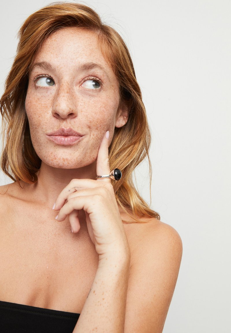 Primer plano de una persona con cabello rojo y pecas, usando un anillo negro con una piedra pulida, contra un fondo neutro.