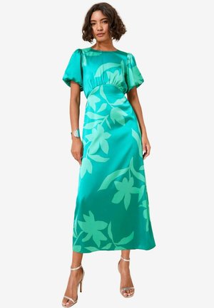 Nainen, jolla on päällään teal-värinen satiinimekko, jossa on suuria kukkakuoseja, puffi-hihat ja avokärkiset korkokengät, seisoo yksinkertaista taustaa vasten.