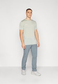 Lichtgroene t-shirt met korte mouwen met een subtiel gradient ontwerp en logo op de borst, gecombineerd met lichtblauwe broek en grijze trailschoenen.
