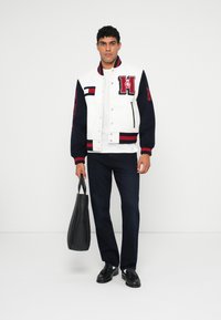 Baltų, jūrų mėlynos ir raudonos spalvų varsity striukė su išsiuvinėtu logotipu, mygtukais snap, kraštiniais rankogaliais, dėvima ant baltos marškinėlės, derinama su jūrų mėlynomis kelnėmis.