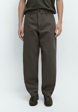 Mann trägt locker sitzende, dunkelbraune Hose mit vorderer Knopfleiste und Reißverschluss, kombiniert mit schwarzen Schnürschuhen, steht vor einem schlichten weißen Hintergrund.