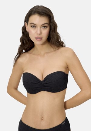 Mujer joven con cabello castaño ondulado, vistiendo un bikini negro texturizado sin tirantes y con pantalones a juego, sobre un fondo claro y liso.