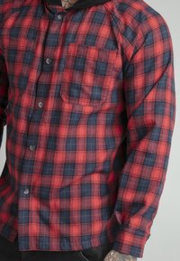 Camicia di flanella a quadri rossi e blu con taschino sul petto, chiusura con bottoni e maniche lunghe. Presenta un tessuto testurizzato e bottoni metallici.