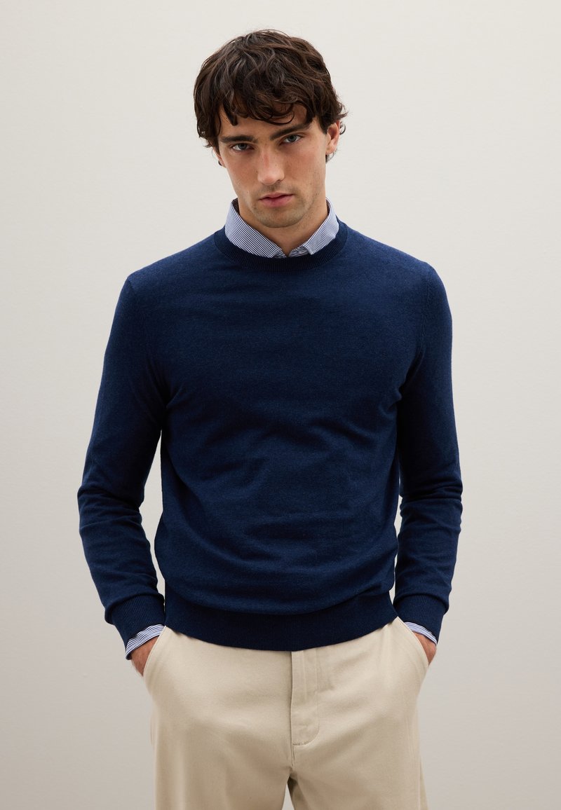 Maglione lavorato a maglia blu navy con scollatura rotonda, maniche lunghe e polsini a coste, indossato sopra una camicia a righe, abbinato a pantaloni beige.