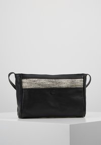 Borsa a mano in pelle nera con una striscia di tessuto bicolore nero e bianco testurizzato e una tasca frontale con chiusura a pulsante, posata su una superficie bianca.