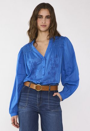 Vrouw die een felblauwe, geborduurde blouse draagt met opgeblazen lange mouwen, gestoken in een blauw spijkerbroek met hoge taille en een bruine riem.