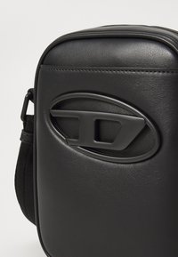 Sac bandoulière en cuir noir avec une forme rectangulaire arrondie et un détail de logo ovale sur le panneau avant.