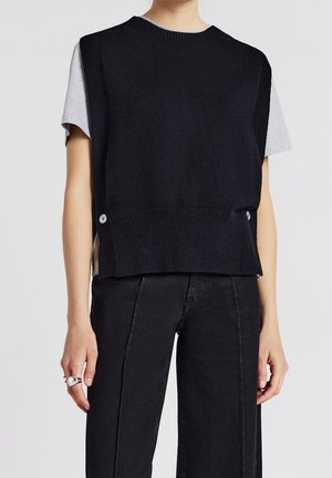 Personne portant un gilet en maille noir sans manches par-dessus une chemise grise à manches courtes, un pantalon noir taille haute et une bague argentée à la main gauche.