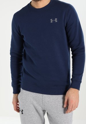 Mann trägt einen dunkelblauen Langarm-Sweatshirt und hellgraue Jogginghose, beide mit sichtbaren Under Armour-Logos.