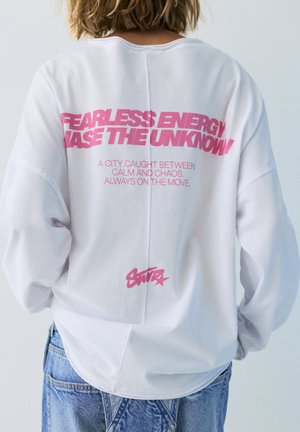 Persoană cu păr blond scurt purtând un hanorac alb cu text roz "FEARLESS ENERGY CHASE THE UNKNOWN" și blugi albaștri.