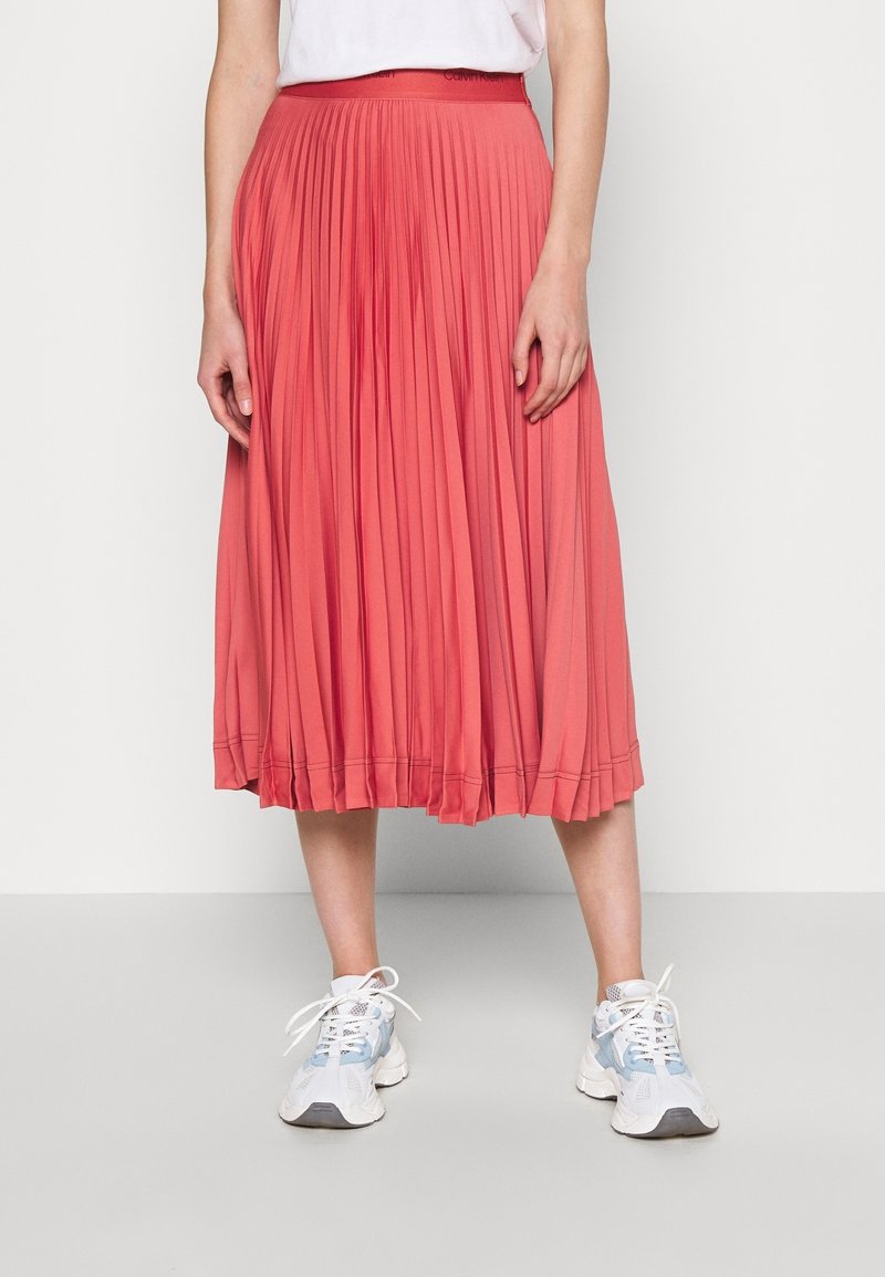 Gonna midi plissettata corallo realizzata in un tessuto morbido, con una vita che presenta il nome del marchio. Abbinata a sneakers bianche e blu.