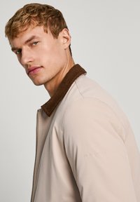 Chaqueta beige clara con un cuello de pana marrón, cierre de cremallera y tela texturizada. Presenta el logotipo grabado de "Pepe Jeans" en la manga.