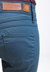 Blauwe denim jeans met een gladde textuur, voorzien van een achterzak en een tan leren patch met ingedrukte branding op de tailleband.