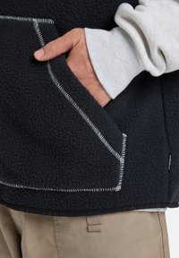 Black fleece vest with white stitched edges, featuring a front pocket, worn over a light gray long-sleeve shirt, with khaki pants visible.
