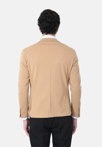 Blazer in tessuto elasticizzato beige con spalle strutturate, apertura centrale e dettagli ai polsi con bottoni; indossato sopra pantaloni neri su uno sfondo bianco.