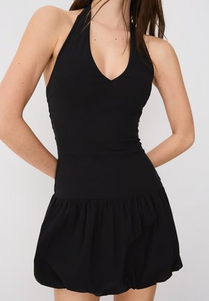 Femme portant une robe noire sans manches avec un décolleté halter, un corsage ajusté et une jupe froncée et évasée sur un fond uni.