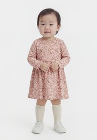 Niña pequeña de pie, vestida con un vestido floral rosa de manga larga, calcetines crema hasta la rodilla y zapatillas blancas, sobre un fondo blanco.