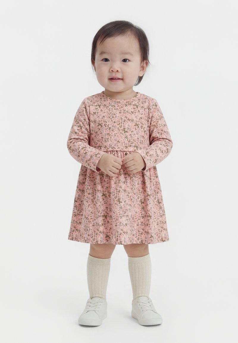 Niña pequeña de pie, vestida con un vestido floral rosa de manga larga, calcetines crema hasta la rodilla y zapatillas blancas, sobre un fondo blanco.